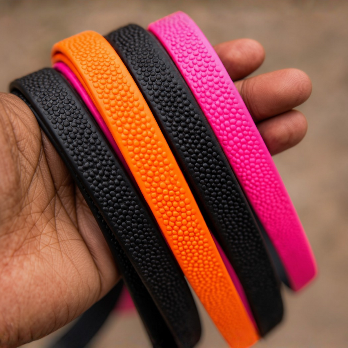 A hand holding four pebble grip Biothane leashes in black, orange and bright pink with raised texture.
